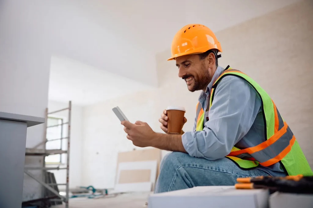Bouwvakker met helm en hesje kijkt op de eigen telefoon in een woning in aanbouw, met een koffiebeker in de hand